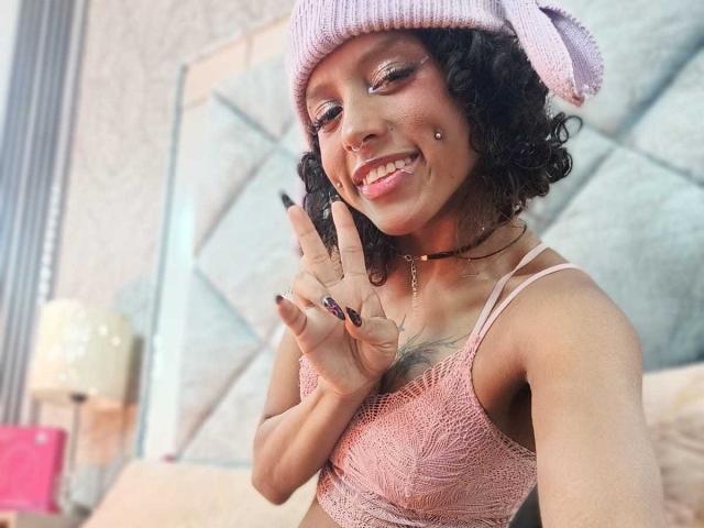 Luna Keller, com cabelos pretos cacheados, usando um gorro rosa de tricô e uma blusa regata rosa de renda, fazendo um sinal de paz, sorrindo. Piercings visíveis no rosto e no nariz. Iluminação interna clara.