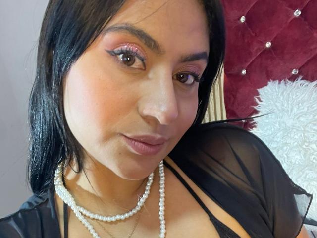 Close-up of DiannaRosse, a Latina woman with long black hair, wearing pink eyeshadow, a pearl necklace, and a black sheer top. She gazes seductively with a slightly parted mouth. Red tufted headboard in background.