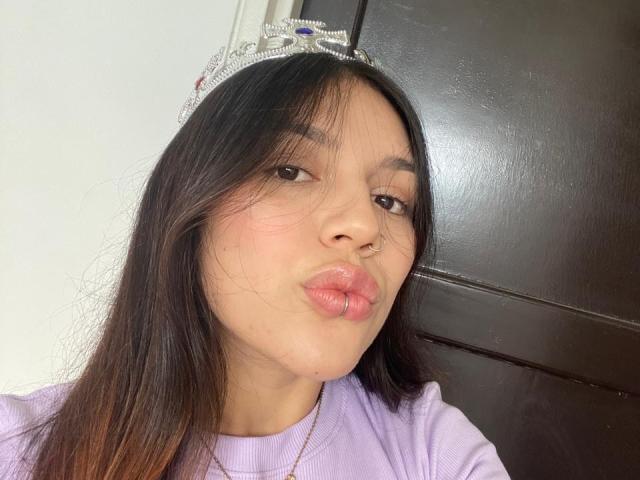 Close-up of SophiaMarx with straight black hair, light brown skin, and glossy pink lips. She's wearing a white shirt, looking sideways with a subtle pout, against a dark background. Fucking sexy.
