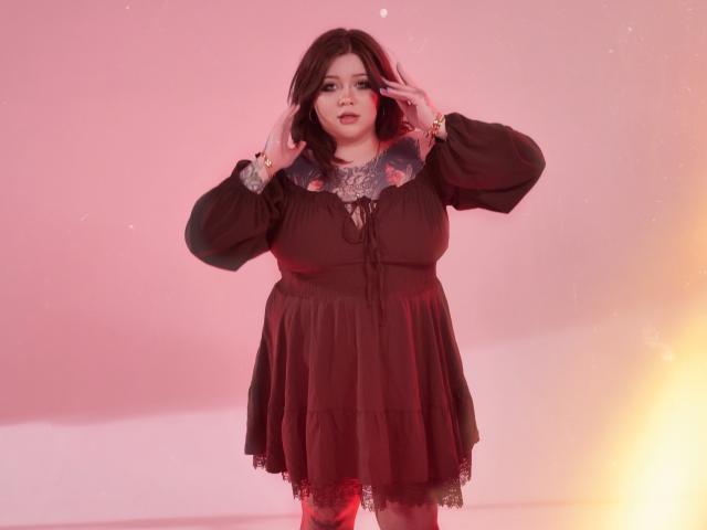 YoureGift, a plus-sized woman with pale skin and dark hair, stands confidently in a black dress with puffed sleeves, hands raised to her head. Pink and orange light glow around her.