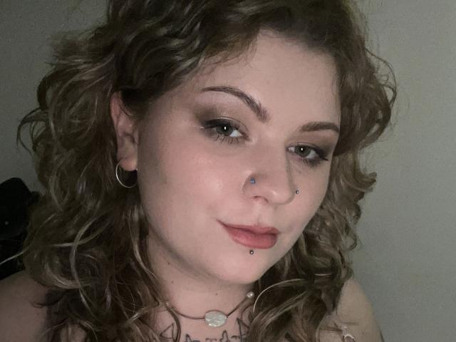 Close-up de AlinaHeiss, uma mulher curvilínea com cabelos castanhos cacheados, pele clara e piercings no nariz e lábio. Ela tem delineador escuro, um sorriso discreto e usa um colar. Incrivelmente sexy..