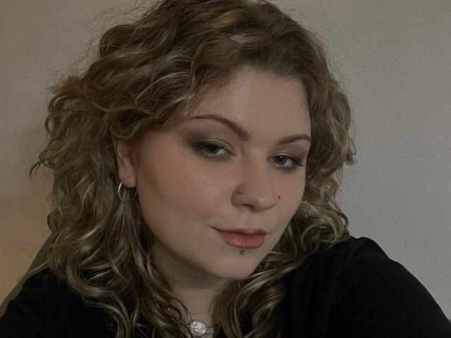 Primer plano de AlinaHeiss, una mujer de piel clara con cabello castaño rizado, que lleva una blusa negra, un pequeño aro en la nariz y aros. aretes. Ella mira hacia abajo con una expresión sutil y seductora..