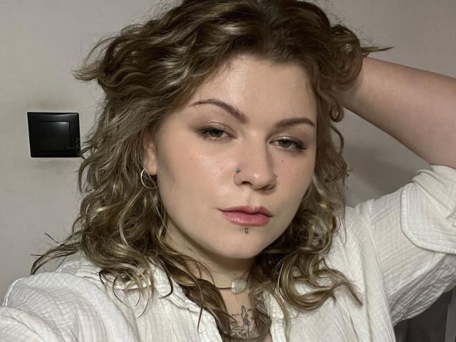 Foto em close-up de AlinaHeiss, uma mulher de pele clara com cabelos castanhos ondulados, vestindo uma camisa branca, com a mão no cabelo, e mostrando o nariz. piercing no nariz, piercing nos lábios, expressão levemente satisfeita, luz suave, fundo cinza..