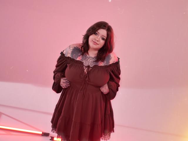 YoureGift, uma mulher plus size com cabelos cacheados escuros, está de pé com confiança em um quarto iluminado por luzes vermelhas. Ela veste um vestido marrom escuro com um decote franzido, que expõe seios grandes. Ela está sorrindo, com as mãos nos quadris..