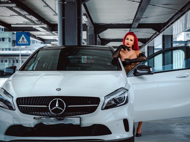 TiffanyBlaize, pelirroja, voluptuosa, vestida con lencería negra, se inclina de forma seductora contra un brillante Mercedes-Benz blanco, en la parte delantera. Puerta abierta, estacionamiento interior luminoso, intensa tensión sexual, entorno urbano moderno, emblema del coche visible..