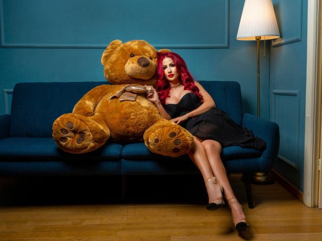TiffanyBlaize with red hair, sitting on a blue couch, hugging a large brown teddy bear. Wearing black dress and heels. Lamp in background, wooden floor, sexual tension.