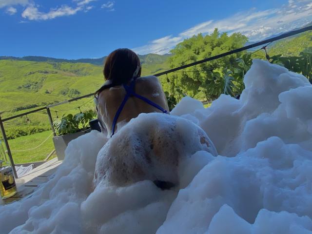 Angeline und Michael sitzen in einer Seifenblasenbadewanne, mit dem Rücken zur Kamera, und zeigen einen nassen, runden Hintern. Sie trägt einen blauen Bikini, im Hintergrund ist eine malerische Berglandschaft zu sehen..