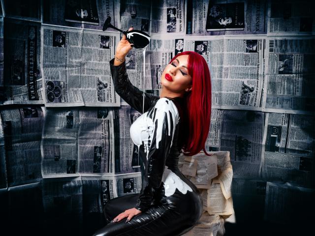 TiffanyBlaize, com cabelo ruivo, vestindo uma roupa de látex preta com detalhes brancos, sentada contra um jornal preto e branco. parede, segurando um chapéu preto, olhos fechados, inclinada para trás, pose sedutora.