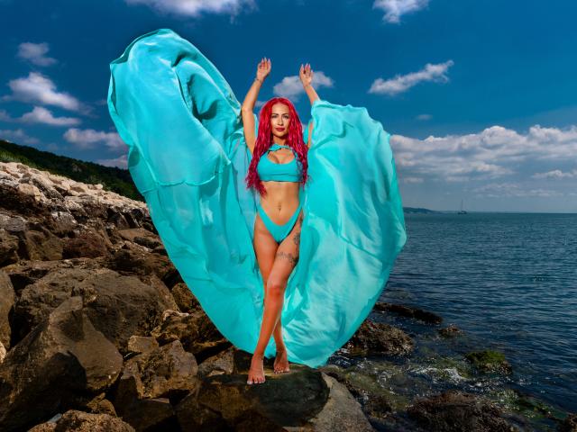 TiffanyBlaize steht an der felsigen Küste, die Arme erhoben, und trägt einen hellblauen Bikini sowie ein durchscheinendes, blaues Tuch, das im Wind weht. hinter ihr. Rotes Haar, verführerische Pose, Ozean und bewölkter Himmel im Hintergrund..