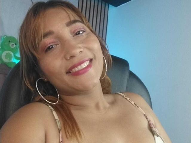 Close-up de MiiaGrey com cabelo castanho, sorrindo, vestindo uma blusa rosa com alças finas, brincos grandes em formato de argola e sombra rosa nos olhos. Brinquedo de pelúcia verde ao fundo. Tensão sexual causada pelo olhar direto..