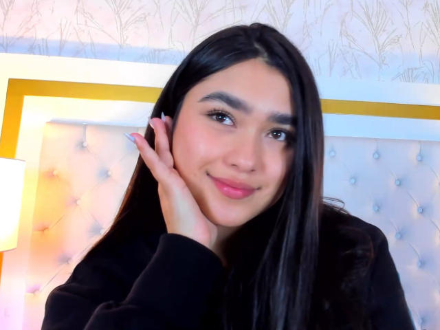 Fotografía de MiaaFerrer, una joven con cabello largo y negro, piel clara y cejas oscuras, sonriendo ligeramente, tocando tocándose la mejilla con la mano, vistiendo una blusa negra, frente a un cabecero brillante y acolchado..