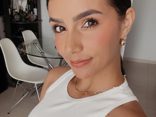 Nahaufnahme von KimmyK, einer Frau mit dunklem Haar, braunen Augen und vollen Augenbrauen, die ein weißes Tanktop, goldene Ohrringe und kette. Im Hintergrund sind weiße Stühle und ein Glastisch zu sehen. Verdammt sexy..