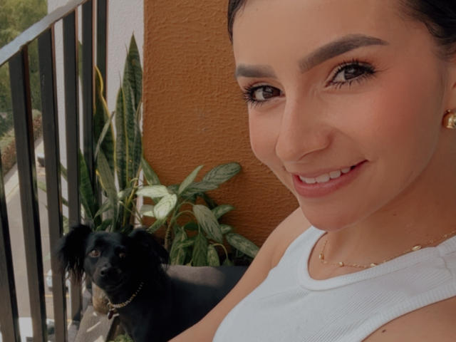 Autoritratto ravvicinato di KimmyK, una donna sorridente con sopracciglia scure, occhi marroni e una canotta bianca, accanto a un cane nero. Pelliccia soffice, foglie verdi sullo sfondo. Tensione sessuale nello sguardo di KimmyK.