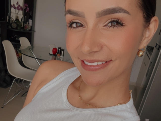 Close-up photo of KimmyK with dark eyebrows, brown eyes, and a smile. She wears a white tank top, gold earrings, and necklace. Background shows modern kitchen with white chairs. Fucking hot.