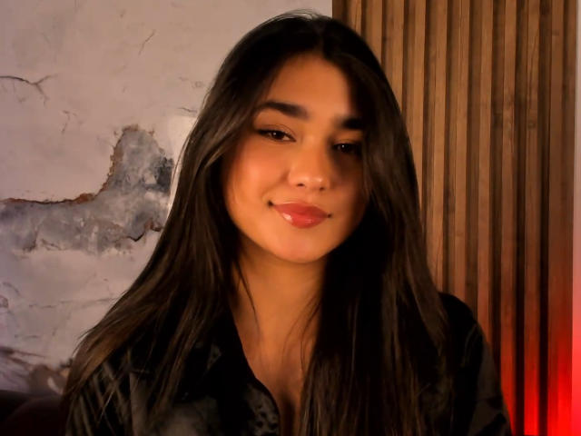 Photograph of MiaaFerrer, a young woman with long black hair, dark eyes, and full lips, wearing a black shirt, standing against a cracked wall and wooden panel, with a red light glow.