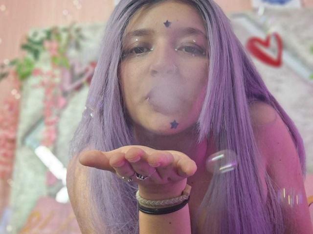 Foto em close-up de CandySwan com cabelos roxos longos, soprando um beijo com uma tatuagem de estrela na testa. Ela está usando uma pulseira e tem um piercing no nariz. O fundo é colorido e desfocado..