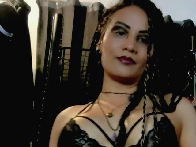 DivaMarie, with dark braided hair and deep red lipstick, wears a black lace top, showing cleavage and strappy details. Background is dark, with shadows. Sexual tension, sultry expression.