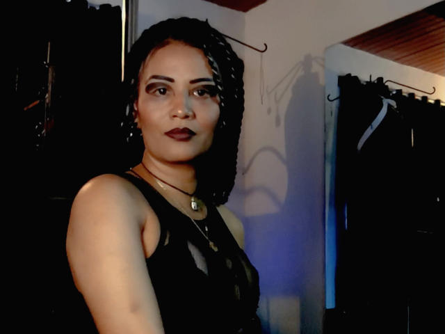 Photograph of DivaMarie with short, curly black hair, wearing a black, sheer top, and dark lipstick. She has a confident, slightly sultry expression, standing in a dimly lit room.