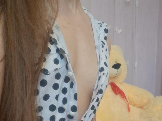 Close-up of Amberia's chest showing a deep neckline with black polka dots on a white dress. Amberia's long brown hair partially covers her. A yellow teddy bear with a red ribbon is on her right.