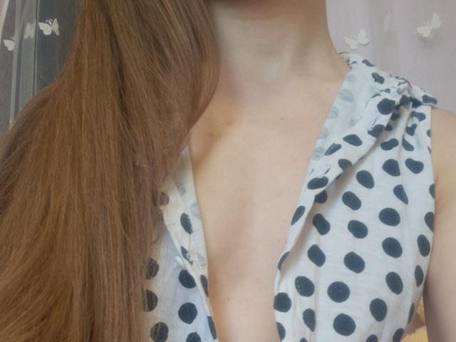 Gros plan de la poitrine et du cou d'Amberia, montrant une chemise blanche à pois avec un décolleté plongeant, mettant en valeur sa poitrine. De longs cheveux bruns tombent sur son épaule. Une lumière naturelle vive.
.