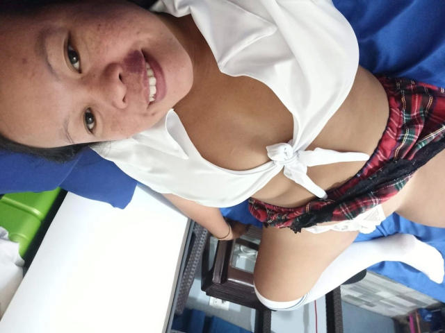 Filipina69-hot, wearing a tied white shirt exposing cleavage and a red plaid mini-skirt, leaning forward with legs apart, smiling directly at the camera. Her cock is partially visible.