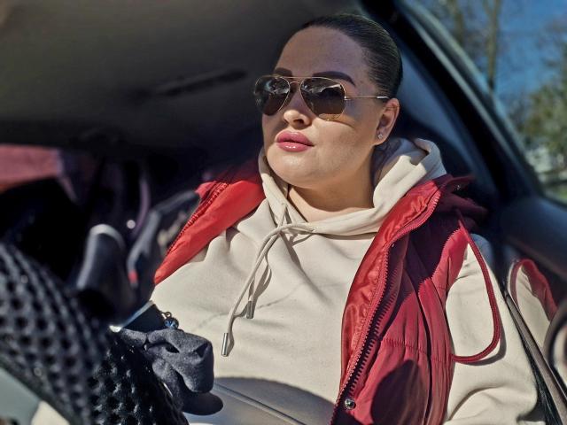 Fotografie von GeorgiaLerox, die in einem Auto sitzt, einen weißen Kapuzenpullover und eine rote Jacke trägt, eine schwarze Sonnenbrille aufhat und etwas in der Hand hält. schwarze Tasche.
das Sonnenlicht betont ihre Gesichtszüge und vermittelt eine sinnliche Ausstrahlung..