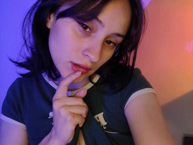 Primer plano de AnnieLamberth, una joven con piel pálida y cabello negro, mordiéndose el dedo de forma seductora. Ella lleva una camisa azul oscuro con detalles blancos. Fondo de luz azul y rosa..