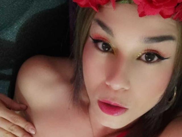 Close-up photo of KimBurton, face slightly tilted, wearing a red flower headband, dramatic black eyeliner, gold eyeshadow, red lipstick, looking up with parted lips, hand near collarbone, sexual tension.