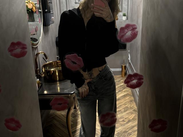 SensualMila stands in a dimly lit room, wearing a black top and tight jeans, hand covering face, mirror reflection showing lipstick prints, gold pot on table, wooden floor. Fucking sexy.