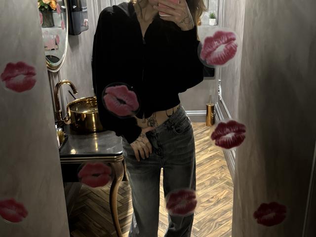Selfie allo specchio di SensualMila, con il viso parzialmente nascosto, che indossa una maglietta nera e jeans attillati, in piedi in un stanza scarsamente illuminata con pavimenti in legno, con sovrapposizioni che ricordano le tracce di rossetto. Mano sul fianco.