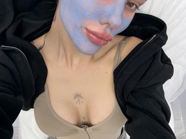 Close-up photo of SensualMila with blue face paint, pouty pink lips, black hoodie, and beige bra. Small sun tattoo below cleavage. Slight tilt, intense gaze, sexy vibe, visible collarbone.