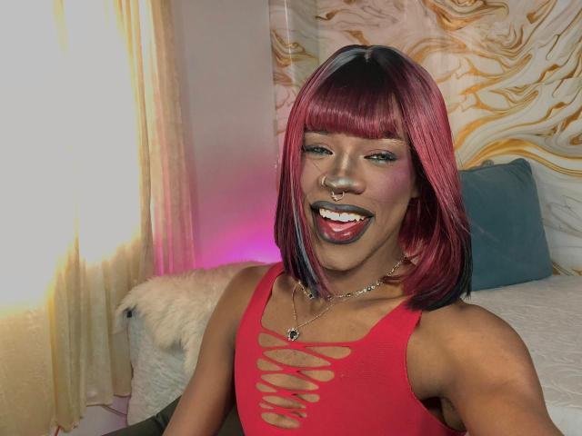 Photograph of EbonyHansNight, smiling with red lipstick, wearing a red tank top with cutout design, nose ring, and necklace. Vibrant marbled wall in background, pink light on left. Fucking sexy.