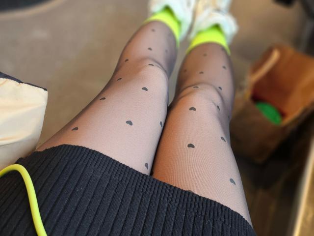 Nahaufnahme der Beine von Red-Rose-hot, die schwarze, durchsichtige Strumpfhosen mit Punkten, einen schwarzen Rock und neon-grüne Schuhe trägt. Saftige Oberschenkel, leichte Textur an den Strumpfhosen, braune Tasche im unscharfen Hintergrund. Verdammt heiß..