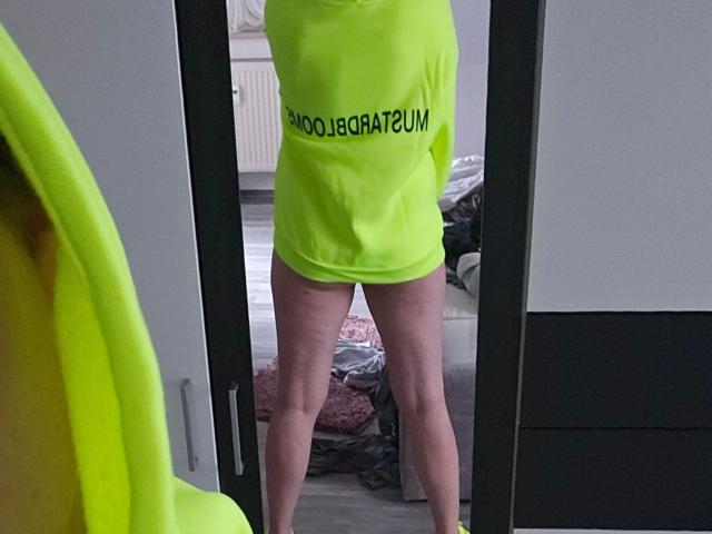 Foto van een reflectie waarop Red-Rose-hot voor een spiegel staat, gekleed in een neongele T-shirt. "GEEN VERPLICHTINGEN. "CUM"-tekst, die blote benen en een glimp van de geslachtsorganen laat zien. Spiegel, deur en rommelige kamer..