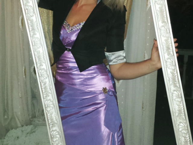 Photograph of Red-Rose-hot wearing a shiny purple dress with a black jacket, holding a glowing white frame. Her right hand rests on the frame, highlighting the dress's sheen.