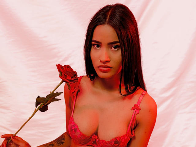 Fotografía de KarolPalacios, una mujer atractiva con cabello largo y negro, sosteniendo una rosa roja en su mano derecha. Ella lleva un sujetador de encaje rosa, que resalta sus grandes senos. La luz roja resalta su expresión sensual..