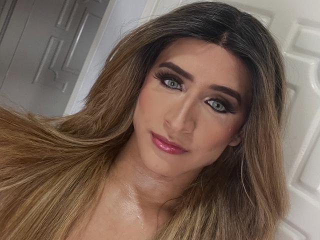 Primer plano de LauraCramz con cabello largo y castaño, ojos azules y lápiz labial rosa. Ella tiene un delineador de ojos negro intenso y máscara de pestañas. Su expresión es ligeramente seductora, con un toque de sonrisa..