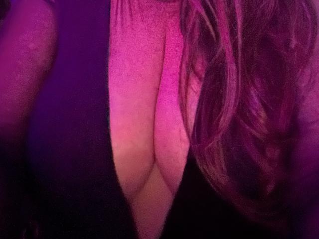 Close-up van de decolleté van BadAngel, waarbij grote, ronde borsten dicht bij elkaar worden weergegeven, met roze en paarse verlichting. Het natte, golvende haar van BadAngel valt naar rechts. De afbeelding heeft een korrelige textuur..