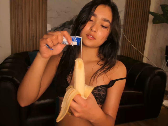 MiaaFerrer with long black hair, wearing a black lace bra, holding a peeled banana and a blue wrapper, licking the tip. She's seductive, eyes half-closed, intense focus on the banana.