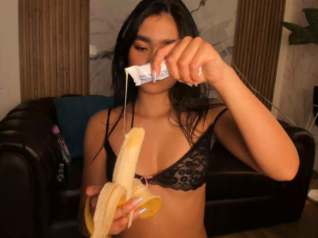 MiaaFerrer, con lunghi capelli neri, indossa un reggiseno di pizzo nero, tiene una banana sbucciata in una mano e un involucro di un preservativo nell'altra. l'altro, seduto su un divano nero. Tensione erotica.