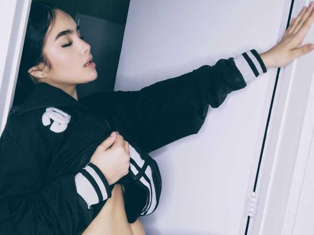 ViolettSweeet se apoya contra una pared blanca, con los ojos cerrados y la boca ligeramente abierta, vistiendo una chaqueta negra con rayas blancas. Brazo derecho levantado, mano izquierda agarrando la chaqueta. Una pose increíblemente intensa y sexy..
