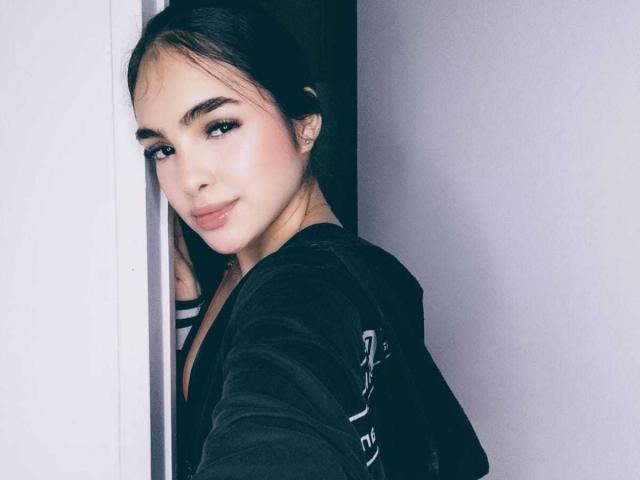 Fotografía de ViolettSweeet con una sonrisa tímida, cabello oscuro recogido en una coleta, vistiendo una blusa negra, apoyada contra una pared blanca. pared, con una luz suave que resaltaba su piel clara y sus labios carnosos. Increíblemente atractivo/a..