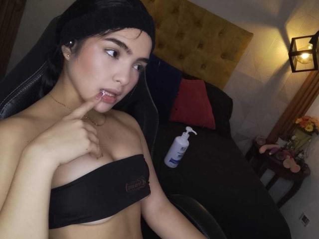ViolettSweeet with black strapless top, finger in mouth, dark hair, seductive gaze. Black hair, light skin, small breasts, gold necklace. Dimly lit room, lamp, black chair, bottle. Sexual tension.