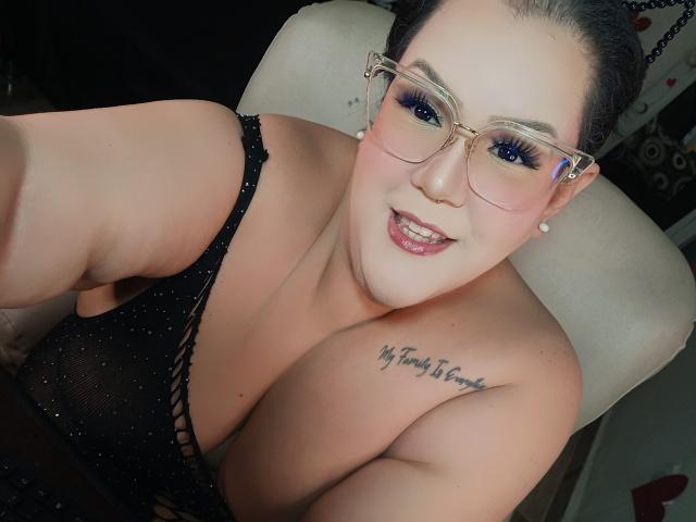 Giianna Morrys, eine kurvige Frau mit großen Brüsten, trägt ein schwarzes, glitzerndes Oberteil und eine Brille. Sie hat ein Tattoo auf ihrem Oberarm. immer an deiner Seite. Sie lächelt verführerisch, die Lippen sind leicht geöffnet..