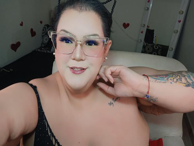 GiiannaMorrys, con i capelli scuri, una fascia con orecchie da gatto e gli occhiali, sorride alla fotocamera, con una mano al collo. Braccio tatuato, top nero con spalline sottili, sedia bianca, adesivi a forma di cuore sulla parete..