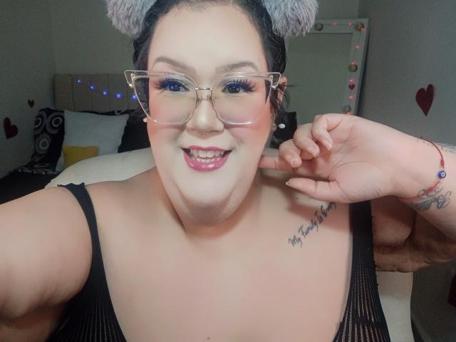 Close-up of GiiannaMorrys, a plus-sized woman with fair skin, black hair in gray buns, wearing clear glasses and a black spaghetti strap top, smiling with her hand near her face. Tattoo on shoulder.