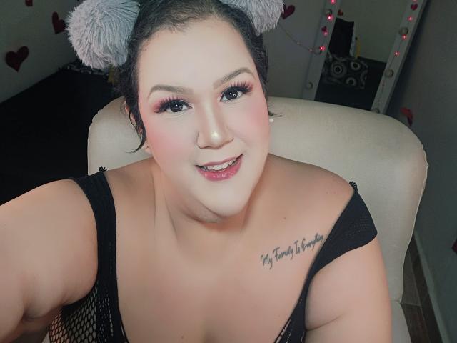 Photograph of GiiannaMorrys with gray furry ear headband, black mesh top, pink eyeshadow, smiling, exposed cleavage, tattoo on right chest, sexy, direct gaze, dimly lit room, red string lights.