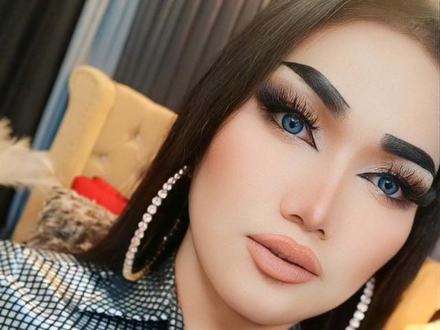 Primer plano de FoxyChelseaTS con ojos azules, cejas negras y gruesas, y labios rosados y carnosos. Ella lleva una chaqueta a cuadros, grandes aretes de perlas y delineador de ojos oscuro. El fondo incluye una silla amarilla. Increíblemente sexy..