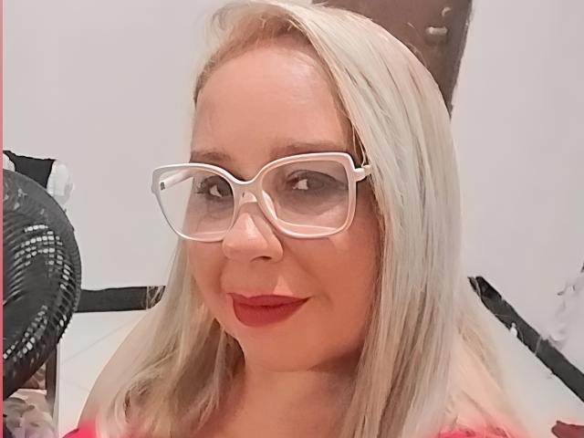 Primer plano de Remitinha con cabello rubio, usando gafas de montura blanca y pintalabios rojo. Tiene una leve sonrisa, una piel radiante y un ligero escote. El fondo incluye un abanico negro..