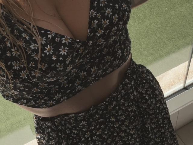 Gros plan de XimenaBella, mettant l'accent sur sa poitrine et sa région abdominale. Elle porte une robe noire à motifs floraux avec un décolleté plongeant, qui révèle une poitrine généreuse et un aperçu de son ventre musclé. Lumière du soleil éclatante.